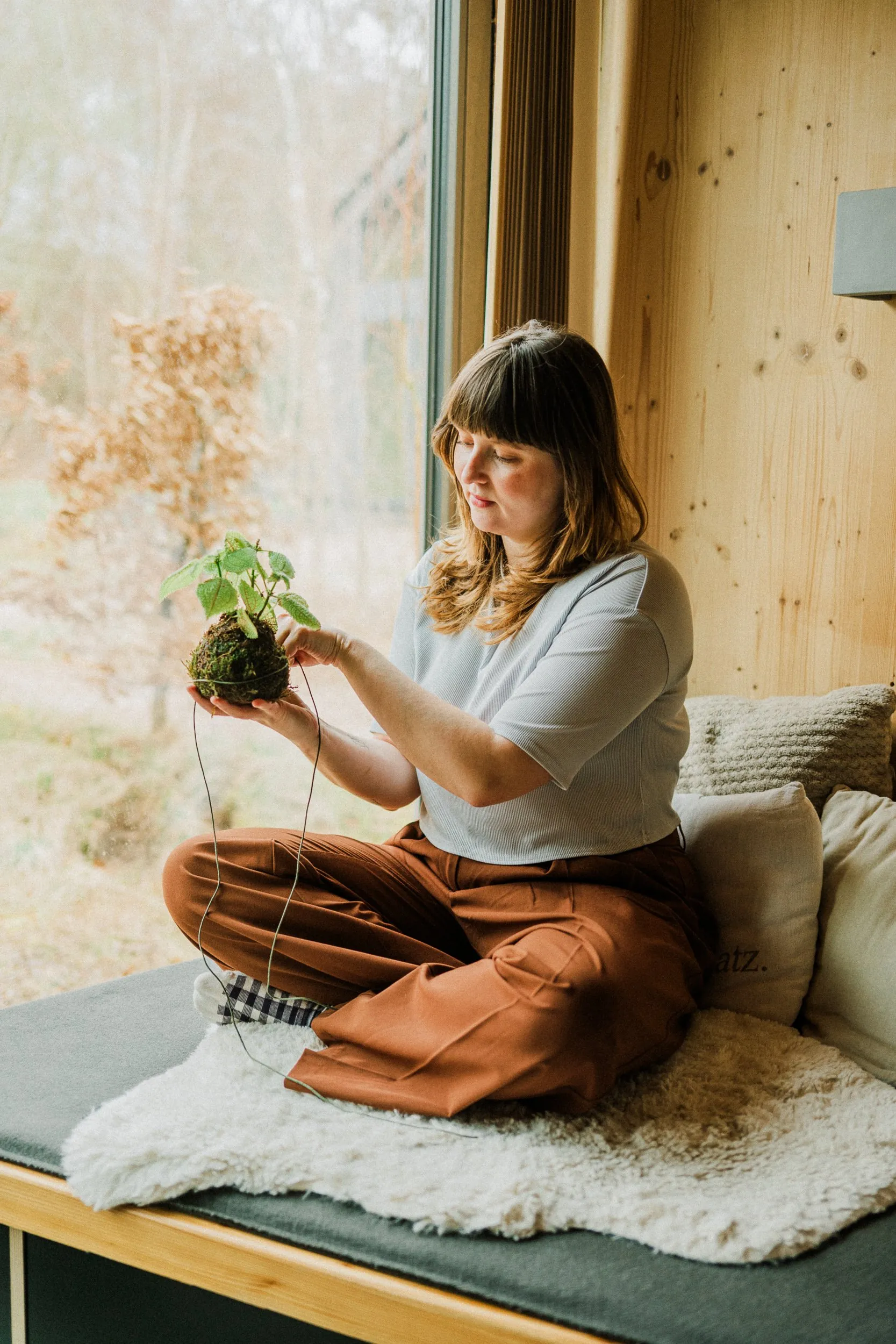 Une femme crée une boule de mousse Kokedama devant la fenêtre panoramique d'un chalet NEUGRAD dans l'Eifel